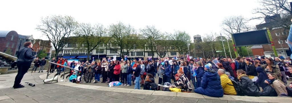 A crowd of people look towards a person to the left of the photography, who is standing at the top of some steps and holding a microphone