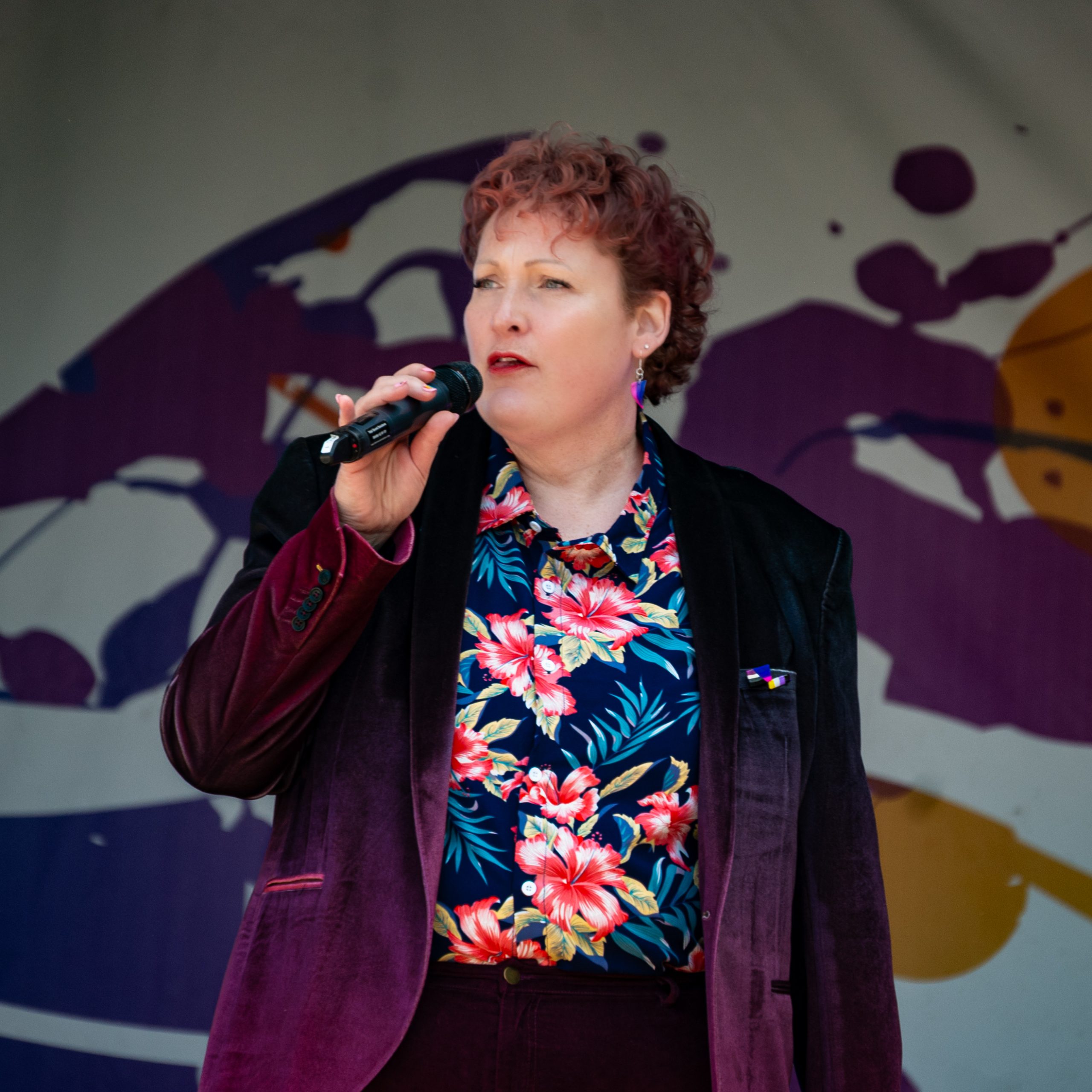 Fair-skinned red-haired person in a floral shirt holds a microphone and talks to a crowd