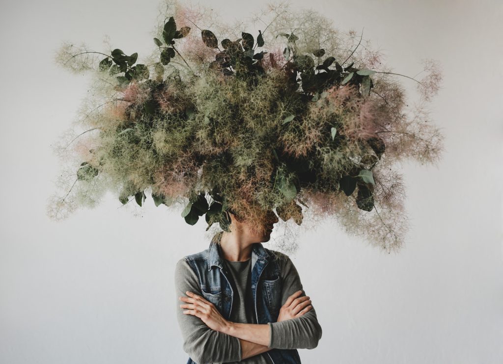 A large decorative bouquet of green leaves, grasses and moss hangs in front of a person's head as though it is coming out of the top of their head. They are standing with their arms crossed over their chest.