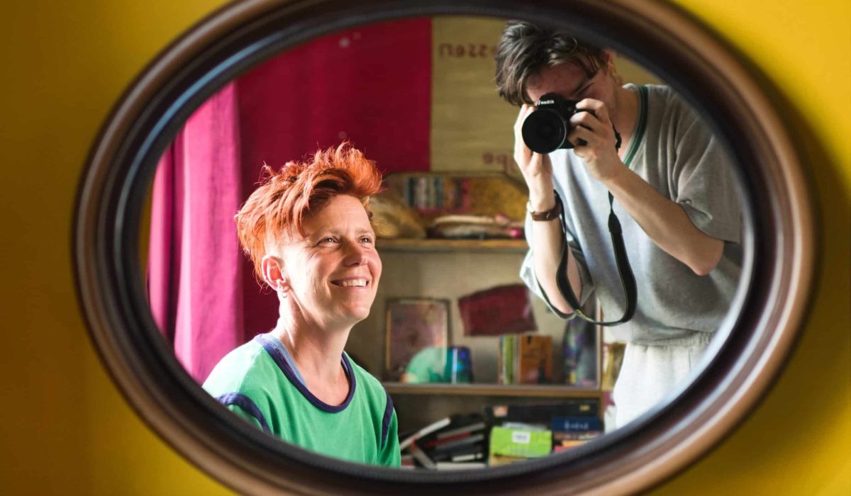 Portret van Mees die in de spiegel kijkt en lacht. In de achtergrond is Lex, de fotograaf, te zien met camera.