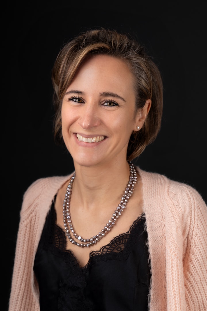 Une femme aux cheveux courts et châtain clair sourit chaleureusement à la caméra dans cette photo professionnelle. Elle porte un cardigan rose clair sur un haut en dentelle noire et un collier de perles, sur un fond noir uni. Cette photo d’entreprise respire la confiance et l’accessibilité | Ce shooting photo portrait professionnel est réalisé par Tribe Photography | Gaëlle Massart Photographe