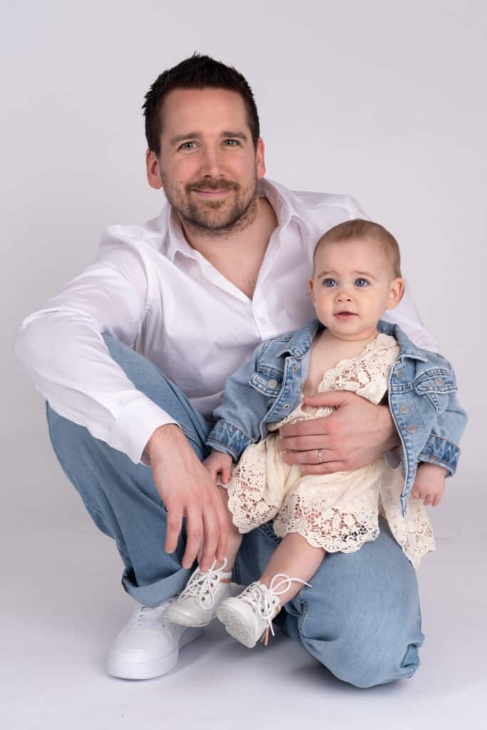 Un homme s'accroupit à côté d'un bébé qu'il tient sur ses genoux lors d'une séance photo de famille. L'homme porte une chemise blanche, un jean bleu clair et des baskets blanches. Le bébé est habillé d'une robe en dentelle blanche et d'une veste en jean bleu clair, avec des petites chaussures blanches. Ils sourient tous les deux à la caméra | Ce shooting photo famille est réalisé par Tribe Photography | Gaëlle Massart Photographe