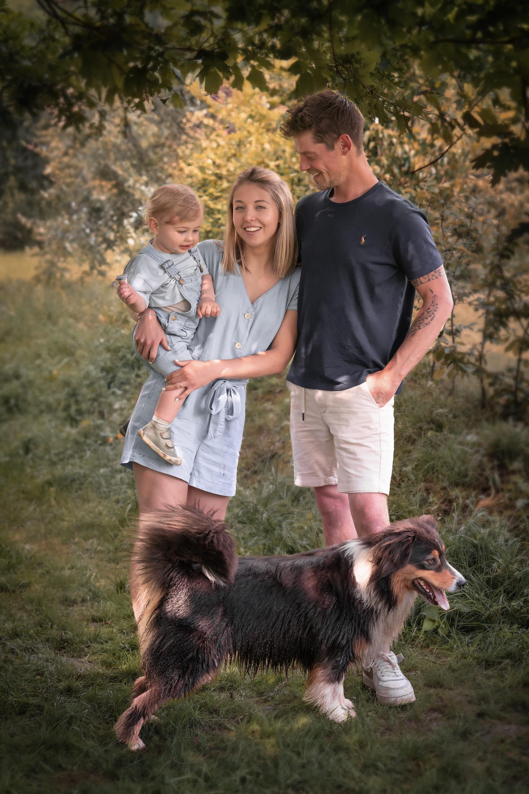 Une famille souriante se tient dehors sur une zone herbeuse, prenant des photos en extérieur. Une femme vêtue d’une robe bleue tient un jeune enfant dans ses bras. À côté d’eux, un homme vêtu d’un t-shirt sombre et d’un short clair les regarde avec tendresse. Devant, un chien à fourrure au manteau foncé et marron pantalon avec la langue sortie. Les arbres sont en arrière-plan | Cette séance photo extérieure est réalisée par Tribe Photography | Gaëlle Massart Photographe