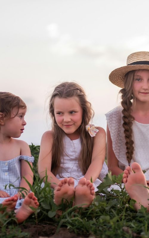 Three sisters in white sundresses are sitting on green meadow. Girls play in blooming field sage