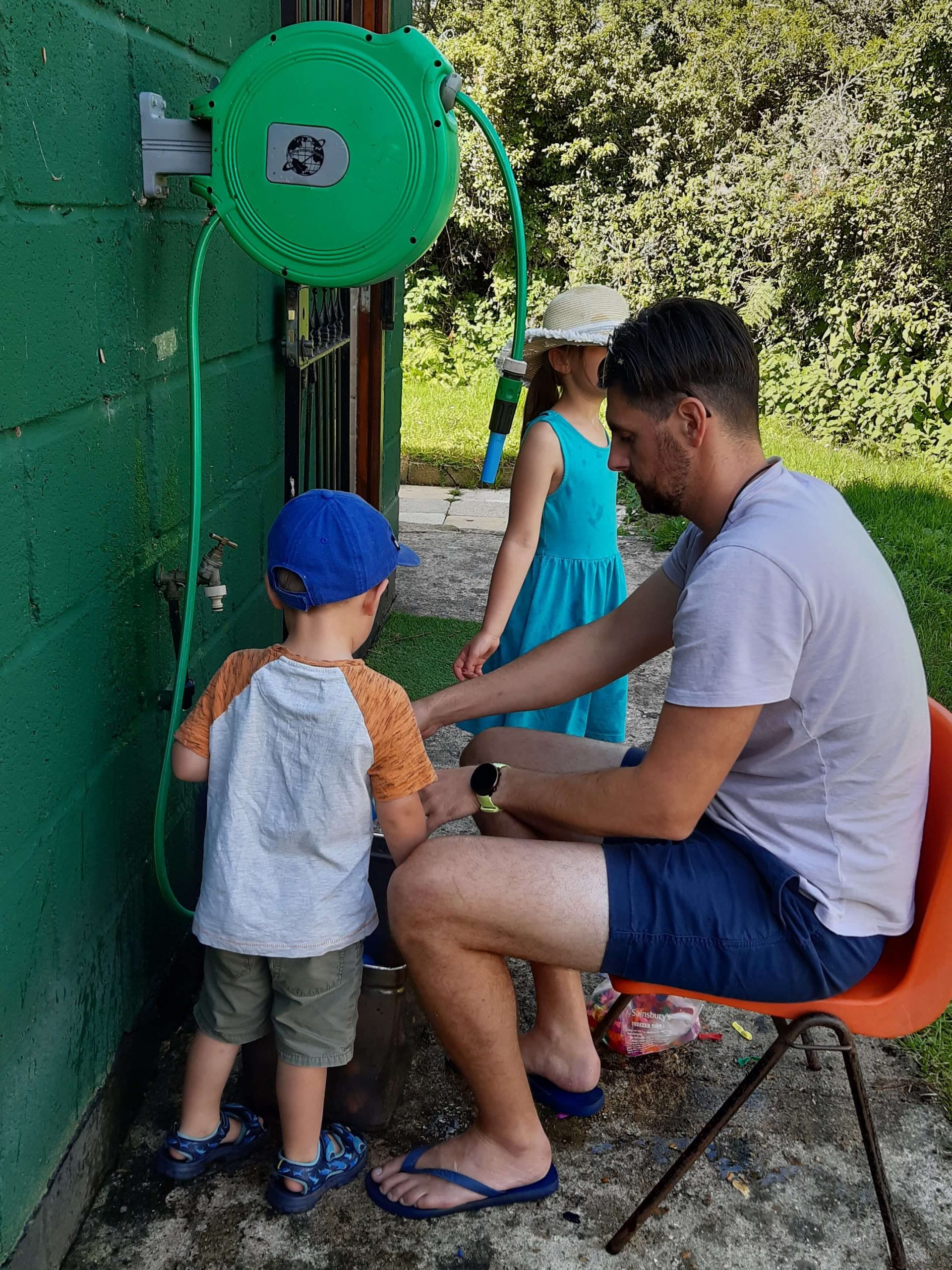 Man sat on chair filling up water balloons from tap. Children watching.