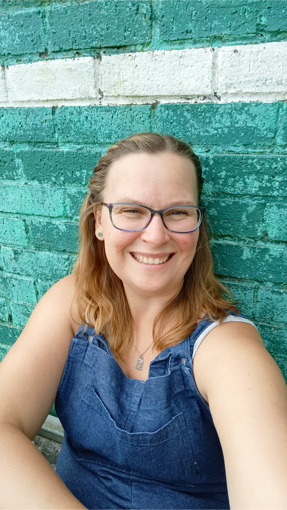 Anna turquoise wall reduced Anna Gilderson in denim dungarees sat against a turquoise brick walll