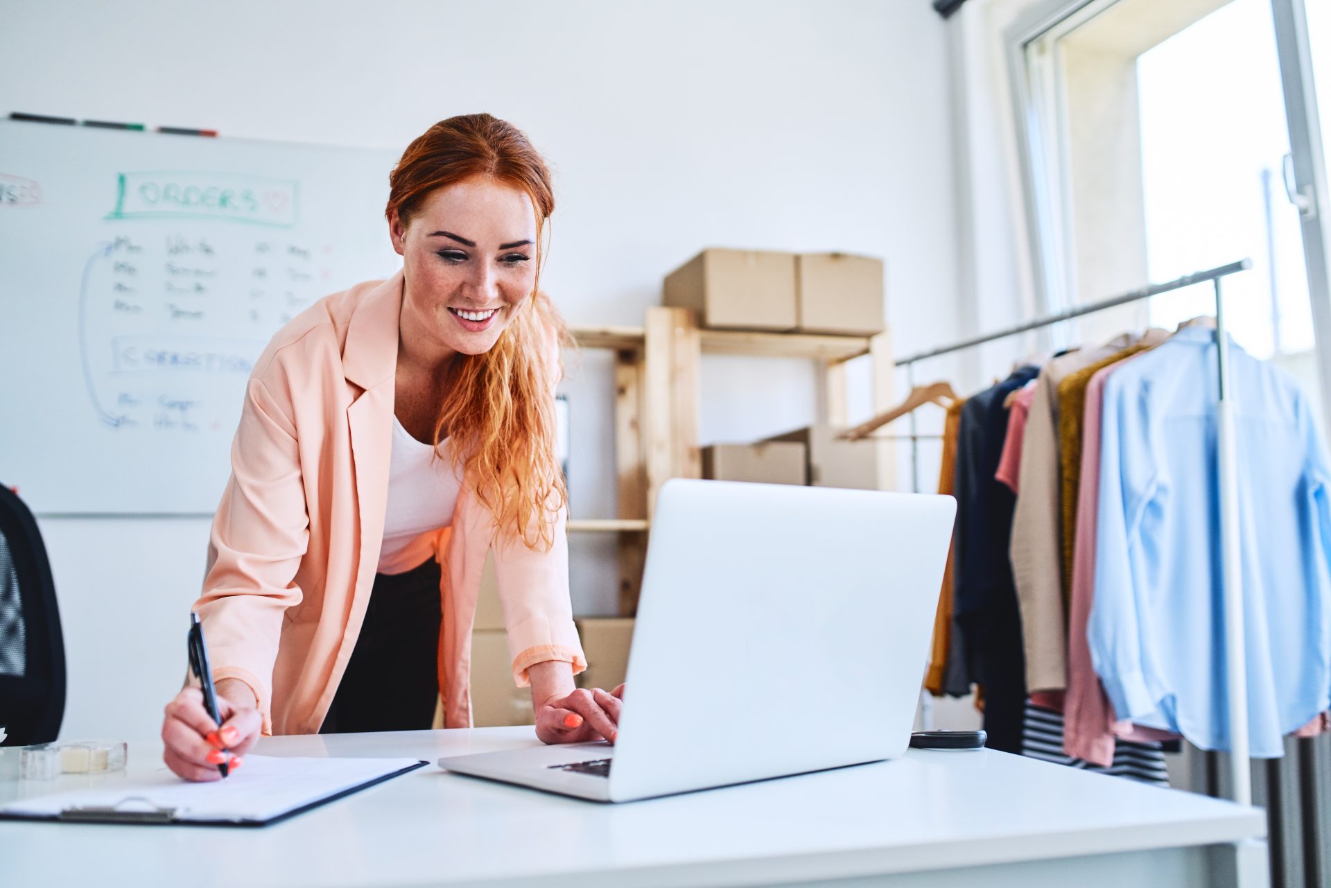 Cheerful female business owner looking at laptop and marking sales and deliveries on clipboard