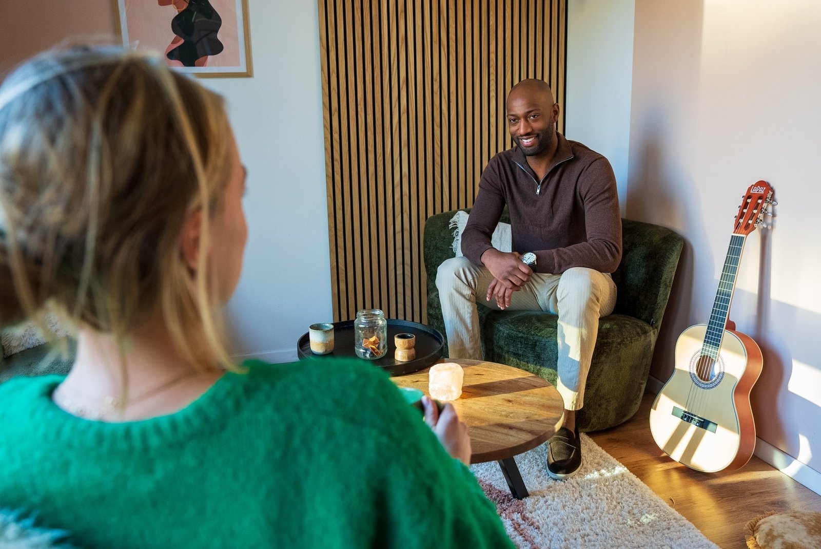 individueel consult bij een bewustzijnscoach in Amsterdam: een man glimlacht terwijl hij in een groene fauteuil zit tegenover een vrouw in een gezellige kamer met gitaar, kaarsen, kleed en kunst aan de muur.
