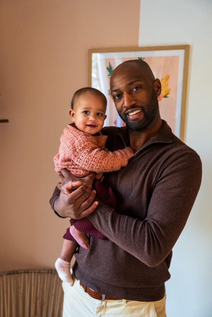 Een bewustzijns coach in Amsterdam dat jouw inzichten geeft en helpt met die belemmerende gedachtes om zo jouw bewustzijn te verruimen, houdt glimlachend een baby vast in zijn armen. De intieme en liefdevolle sfeer weerspiegelt verbinding en balans, passend bij zijn coachingsaanpak. Op de achtergrond is een zachte roze muur met een kleurrijke kunstprint te zien.