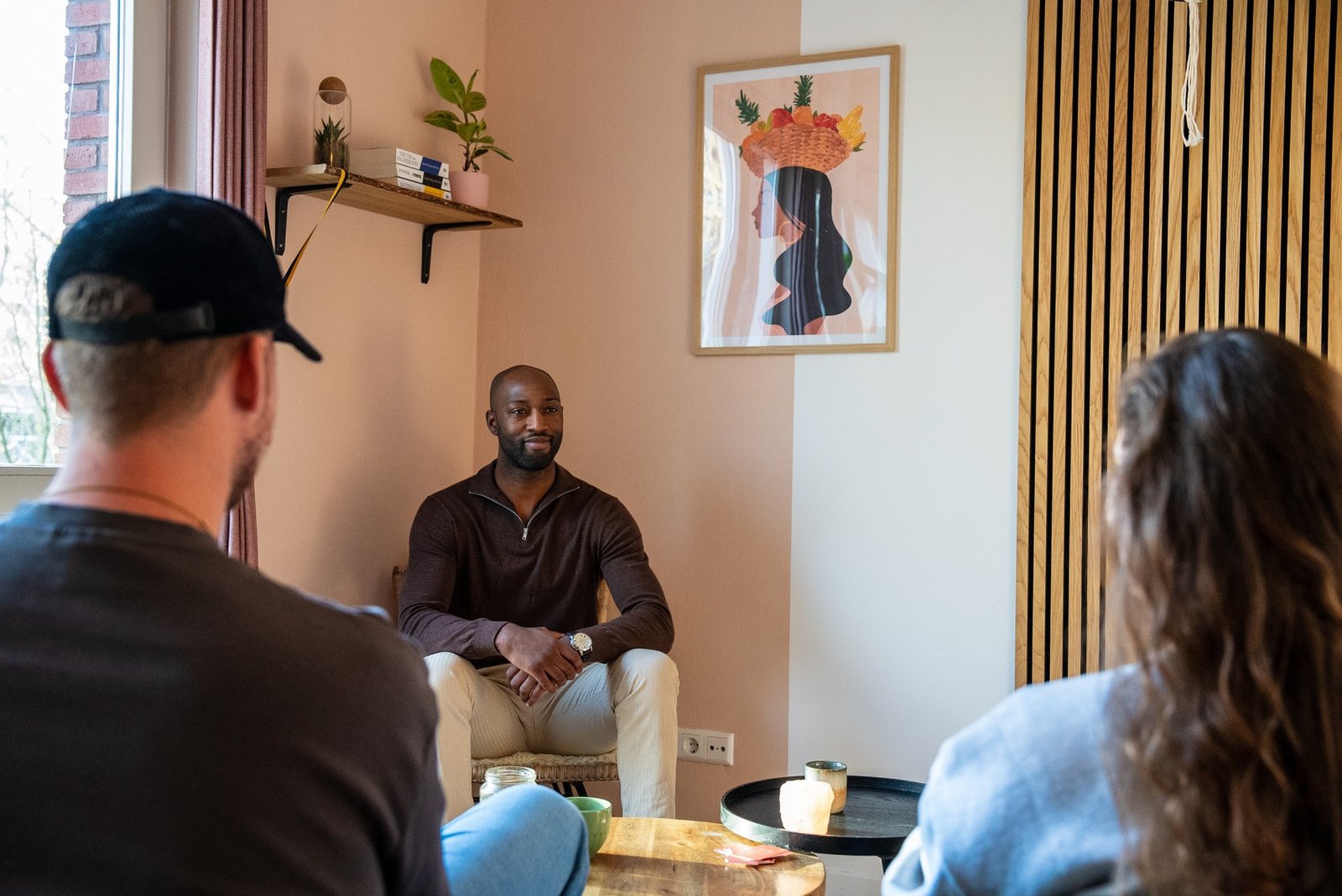 Een bewustzijnscoach voor relaties zit in een gezellige, zonovergoten kamer met lichtroze muren, planten en een abstract kunstwerk, terwijl hij een ontspannen gesprek voert met twee cliënten.