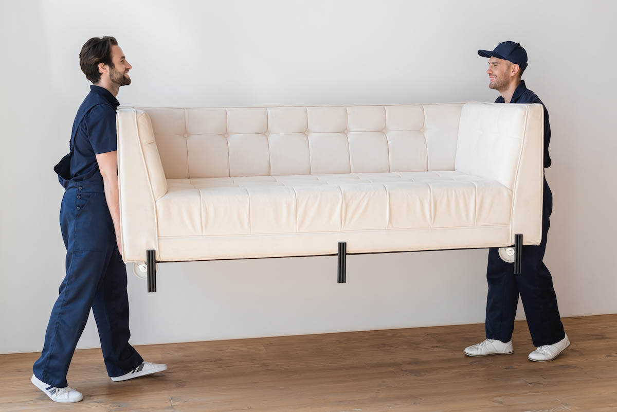 side view of smiling workers lifting white sofa in apartment