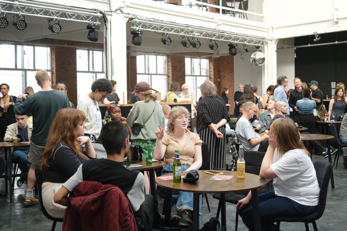 Inside a warehouse space, people sit and stand in small groups chatting.