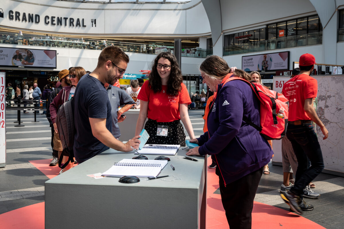 A volunteer wearing a red Key to the City shirt stands in the middle of Birmingham New Street station. They are standing between two members of the public and are smiling.