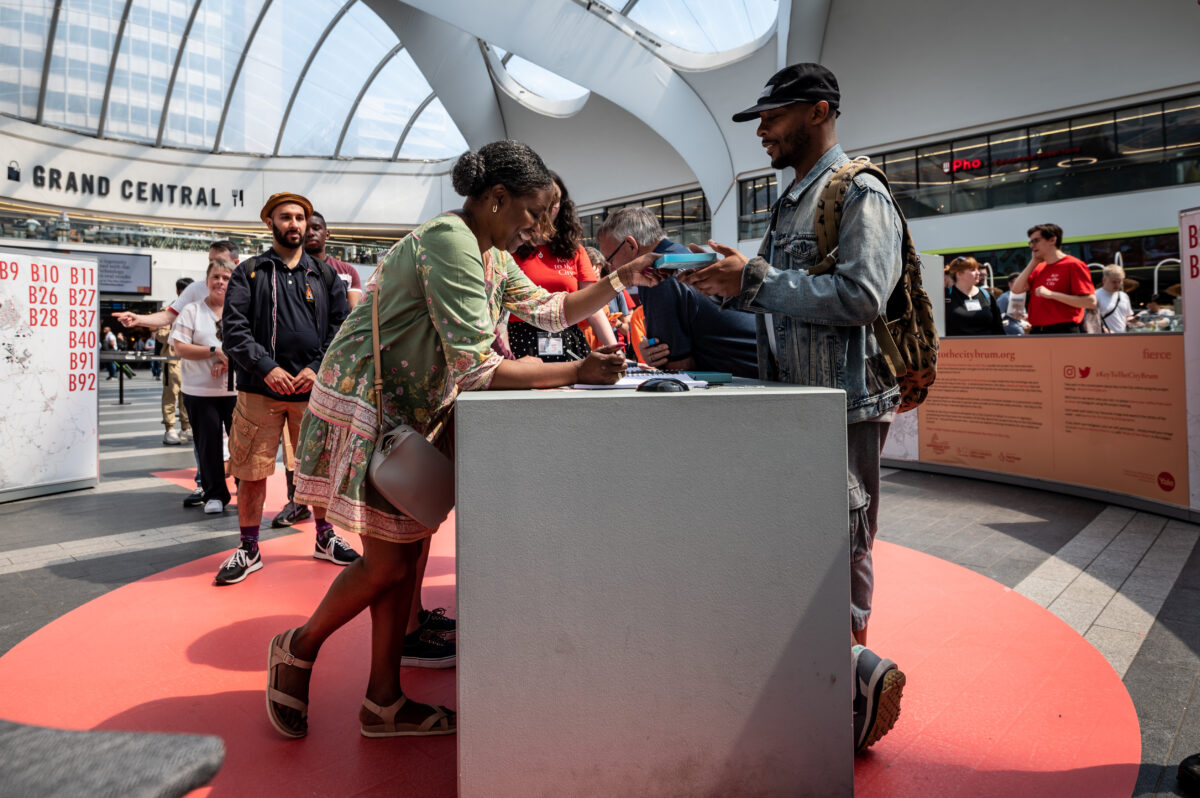 Two people stand across a desk from each other. One hands the other a small blue box as they sign a document on the desk.