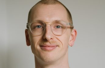 Ethan, in front of a grey wall, is wearing clear glasses and looking into the camera, smiling.