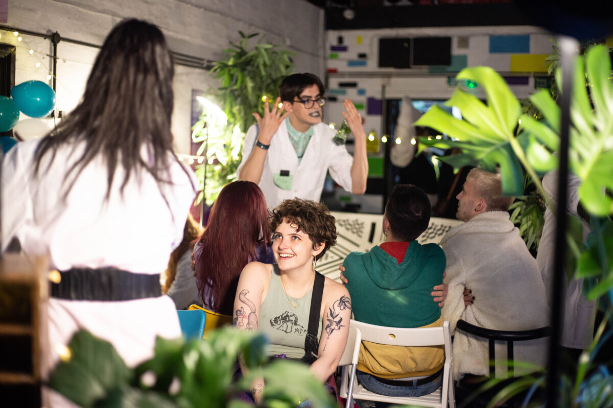 A person with short curly brown hair and tattoos, wearing a vest top, sits on a chair smiling and looking up at a person with their back to camera. The standing person wears a white lab coat and haas long dark hair. In the background three people sit on chairs facing away from camera, they are looking up at a person in a white lab coat, who is gesturing animatedly with their hands. There are several plants in the foreground and background of the image.