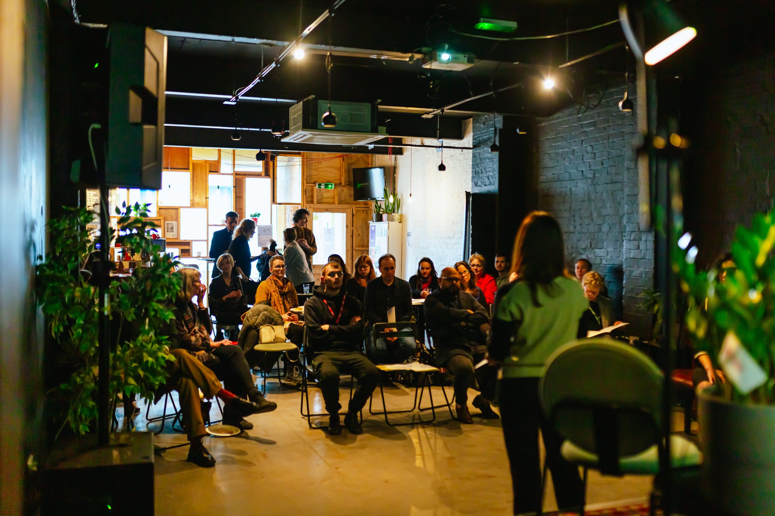 An audience is seated, attentively listening to a speaker whose back is to the camera.