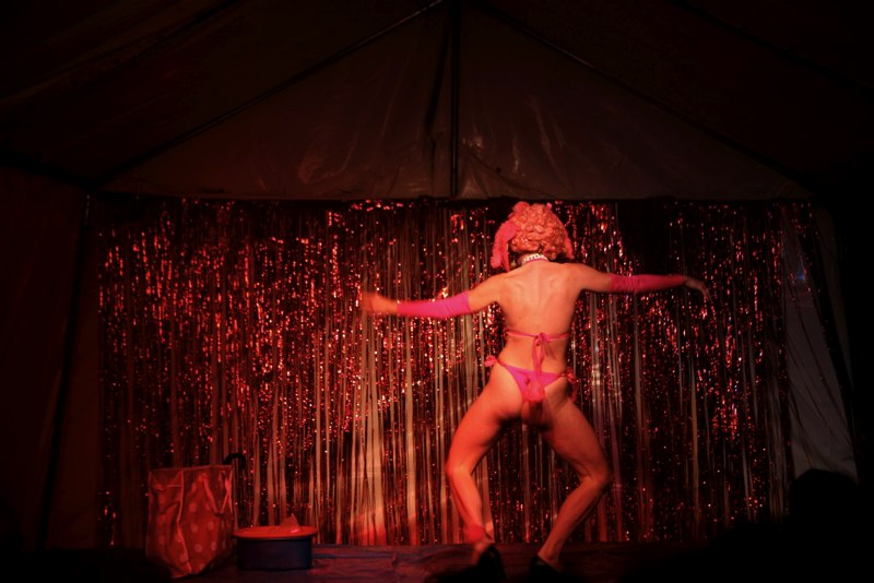 A person wearing a bikini and heels dancing on a stage. They are back to camera and the stage backdrop is glittery ribbon.