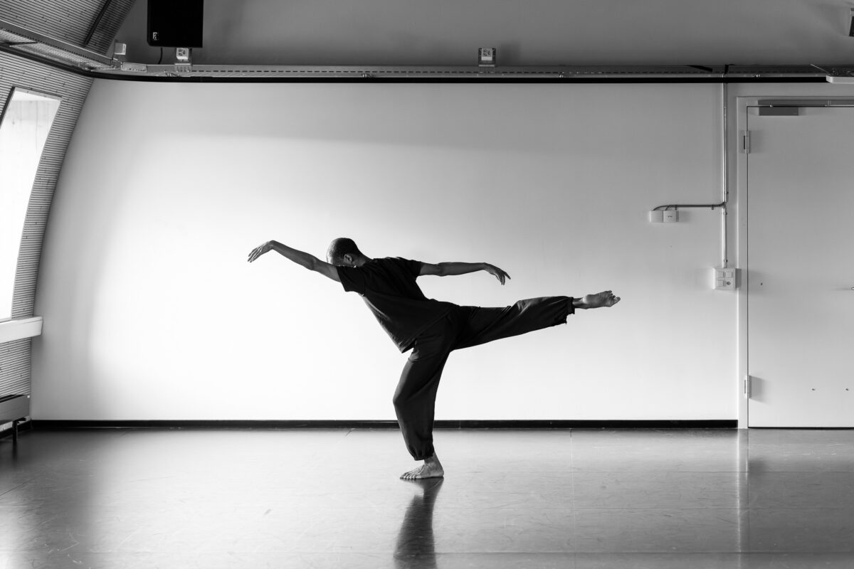 A person wearing a t-shirt and jogging bottoms is doing an arabesque in front of a white wall. The image is in black and white.