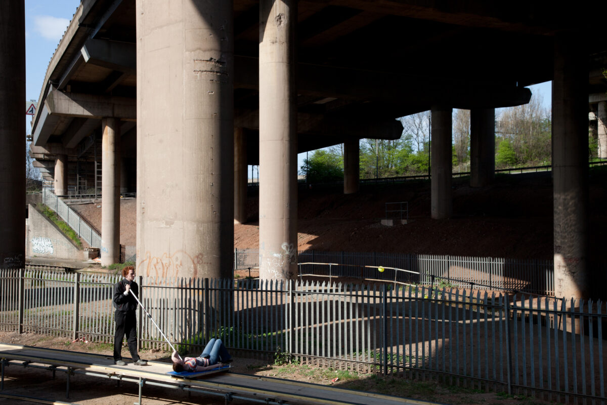 A person lays on their back looking up to the sky, they are being pushed along by a second person. In the background you can see a motorway flyover bridge.