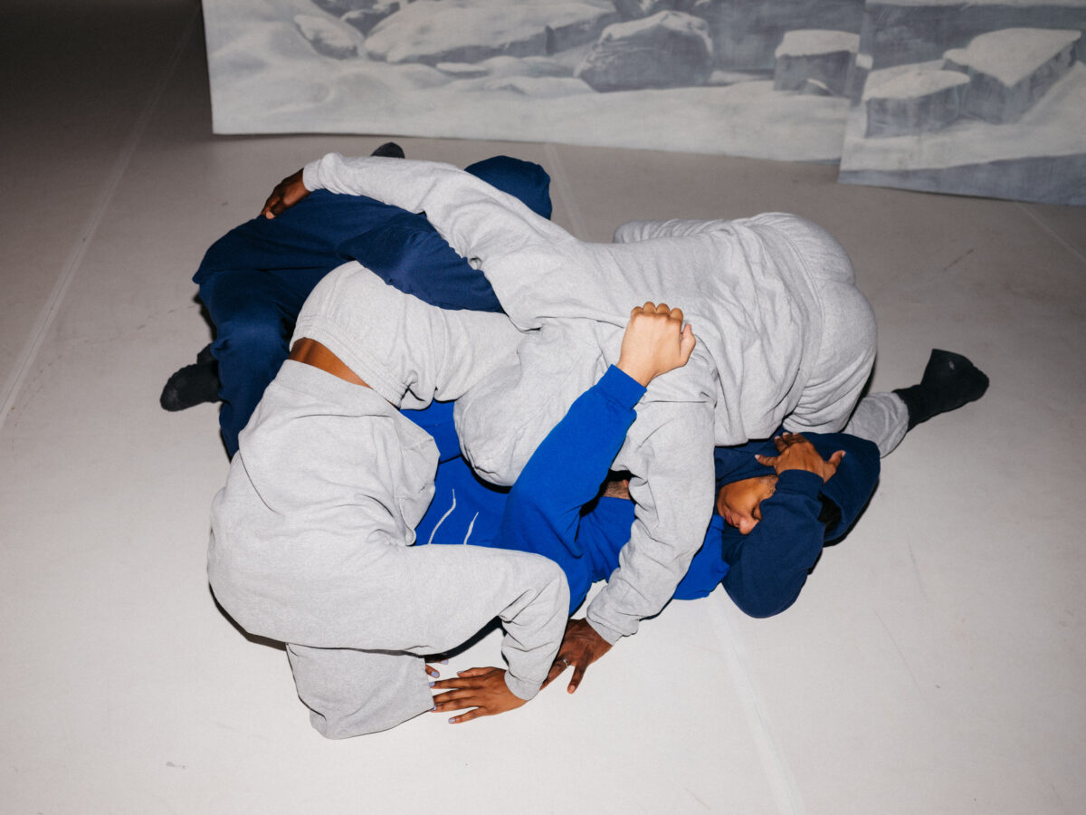 5 bodies are wrapped around each other in a pile. They are wearing grey and blue tracksuits. There is a backdrop of snowy mountains behind them.