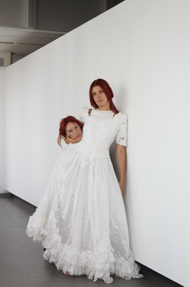 A person with red hair is wearing a wedding dress leaning on a white wall. They have a bodiless head under their left arm, showing a different person’s face, also with red head.