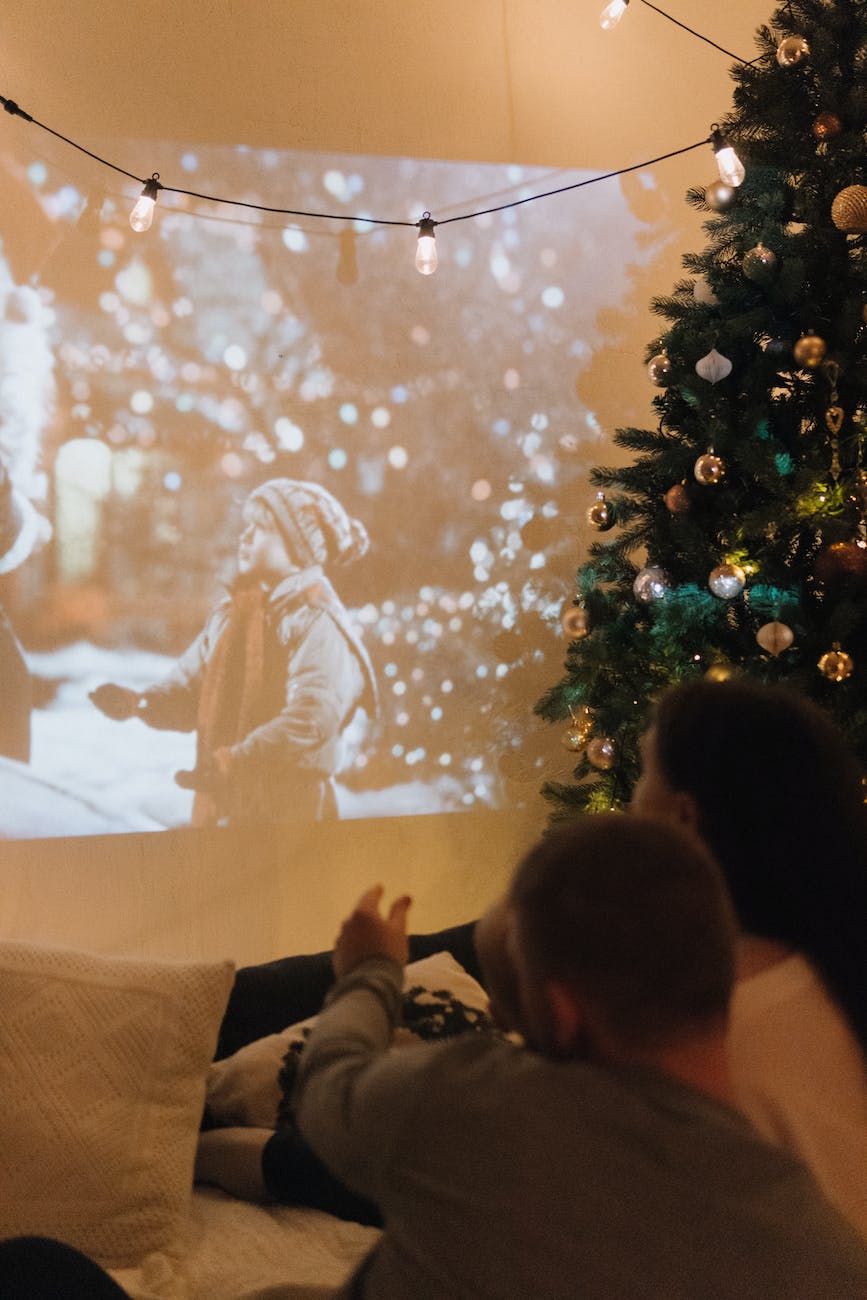 a couple watching a movie displayed on a wall from a projector next to a christmas tree