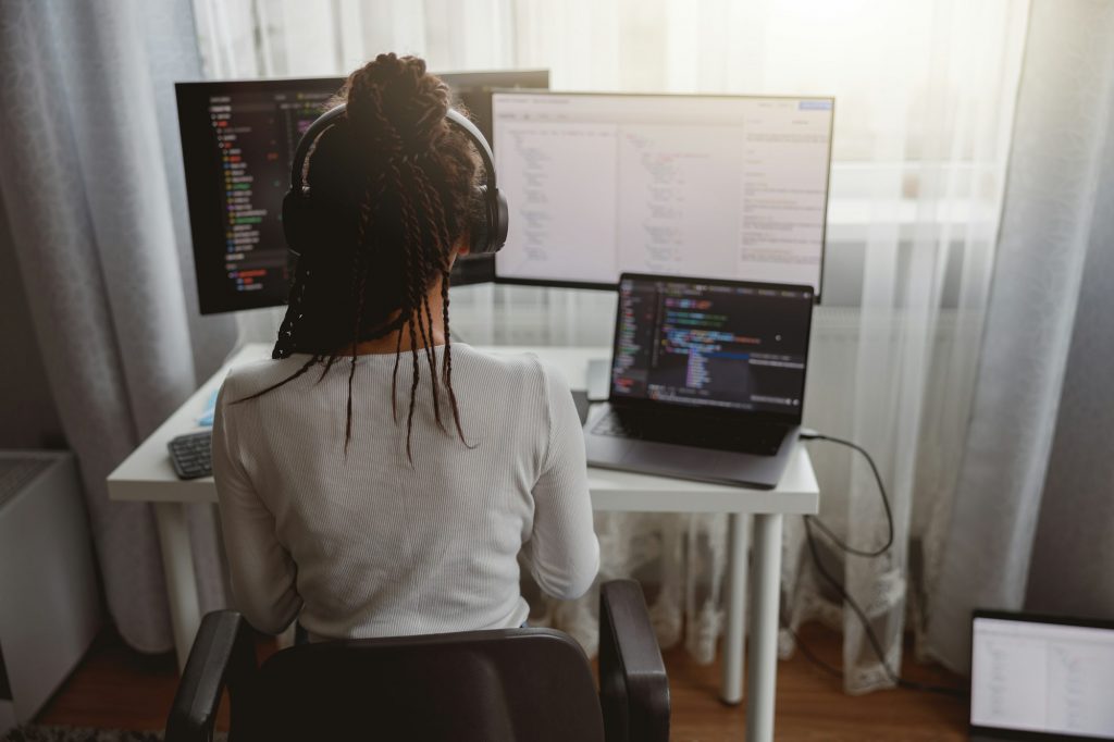 Rear of woman with box braids sitting at table coding on laptop looking at compter