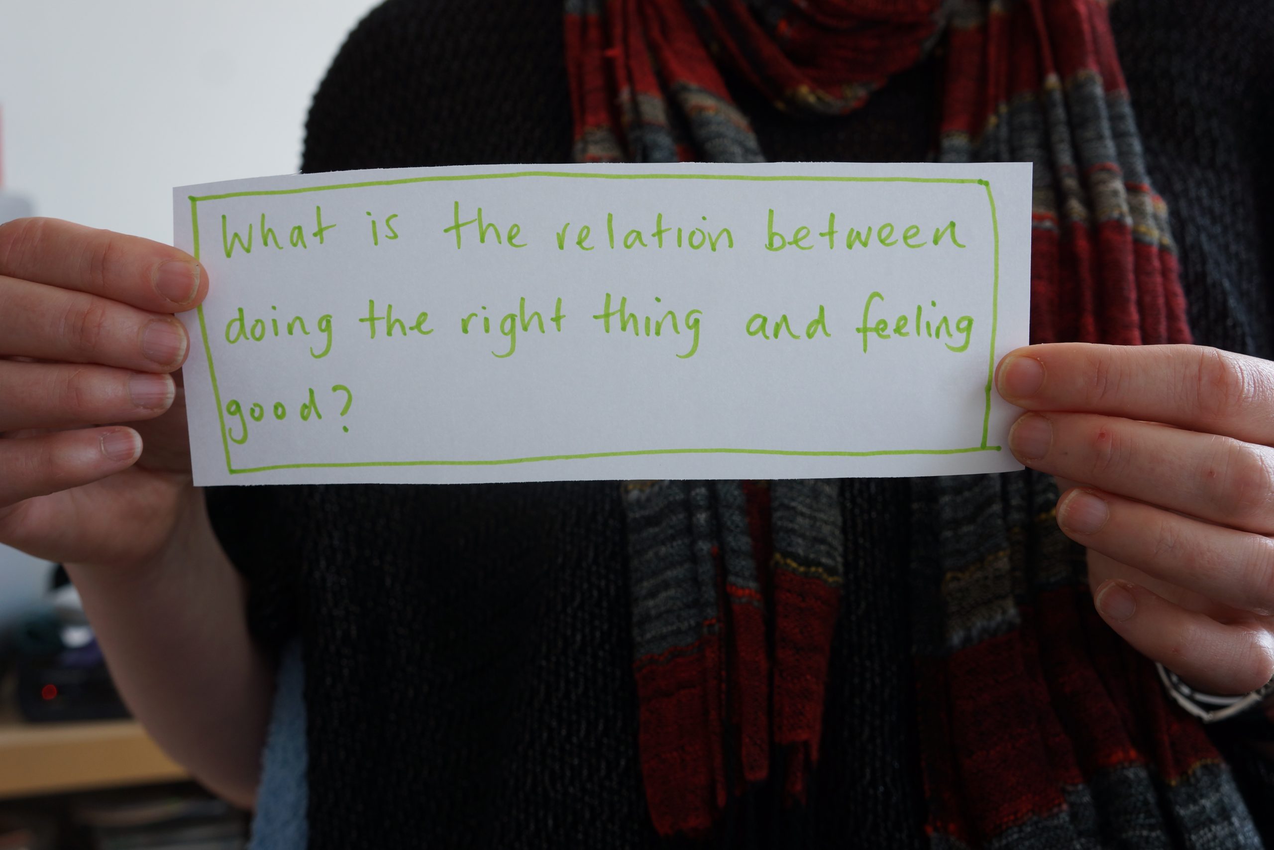 Close up image of hands holding a piece of paper that says in handwritten green text "What is the relation between doing the right thing and feeling good?"