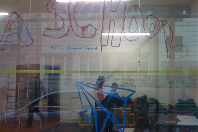 A shop window with large red handwritten words that read "A SCHOOL" and smaller letters in blue that read "Part of Wunderbar Festival" alongside a blue handdrawn image of a paper airplane. Three people can be seen sitting and standing inside the shop.