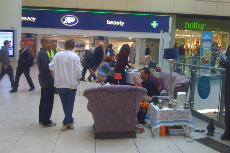 The interior of a shopping mall. People are standing, sitting and chatting by some incongruous old sofas that are outside of a Boots shop and a Hotter shop