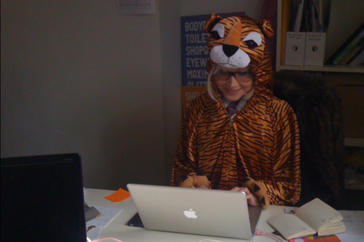 A blond haired white woman with glasses is working on a laptop in an ofice. She is wearing an all-in-one cartoon tiger costume.