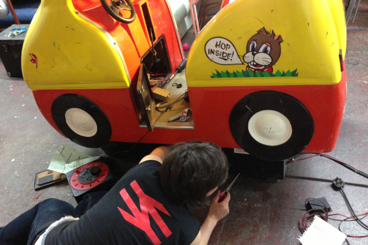 A person is laying on the floor with their back to the camera. They are fixing a coin-operated kids yellow and red toy car ride. A cartoon animal on the car has a speech bubble that says "Hop inside"