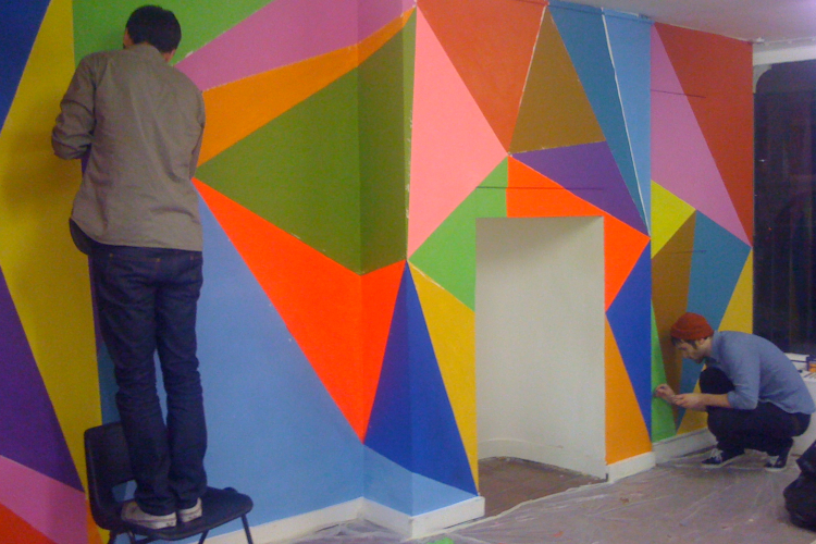 Two men are drawing on a wall. One stands on a chair with their back to camera and one crouches. The wall has a bright multicoloured mural made of large triangles of various sizes and orientations