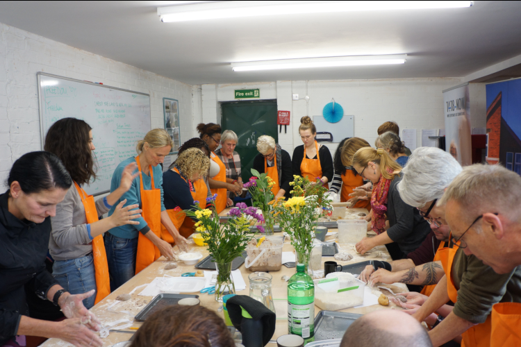 A group of about 20 people stand around a central long table in a small classroom. They are wearing orange aprons. They are concentrating on making things on the table with their hands. The table is decorated with wild flowers in vases.
