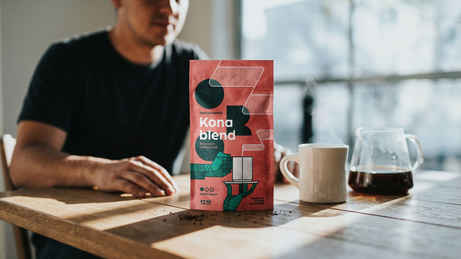 ORO Kona Blend coffee packaging displayed on a wooden table beside a coffee cup and glass carafe in a warm lifestyle setting. Packaging design by Bart De Keyzer featuring geometric illustration, bold typography, and contemporary coffee branding.