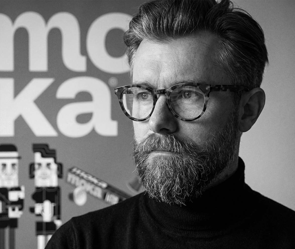 Black-and-white editorial portrait of freelance graphic designer and illustrator Bart De Keyzer (3rd Floor) wearing glasses and a black turtleneck in a creative studio environment with typography artwork in the background.