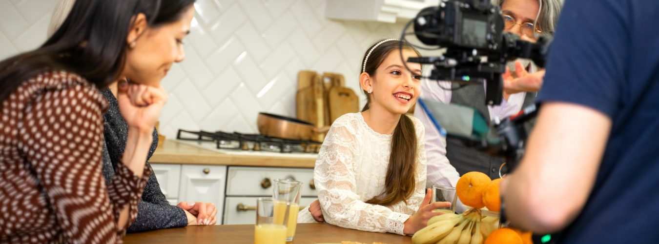 Big multigenerational family having photo or video shoot when cute smiling little girl daughter posing for camera sitting at kitchen table with grandparents and mother. Birthday celebration concept