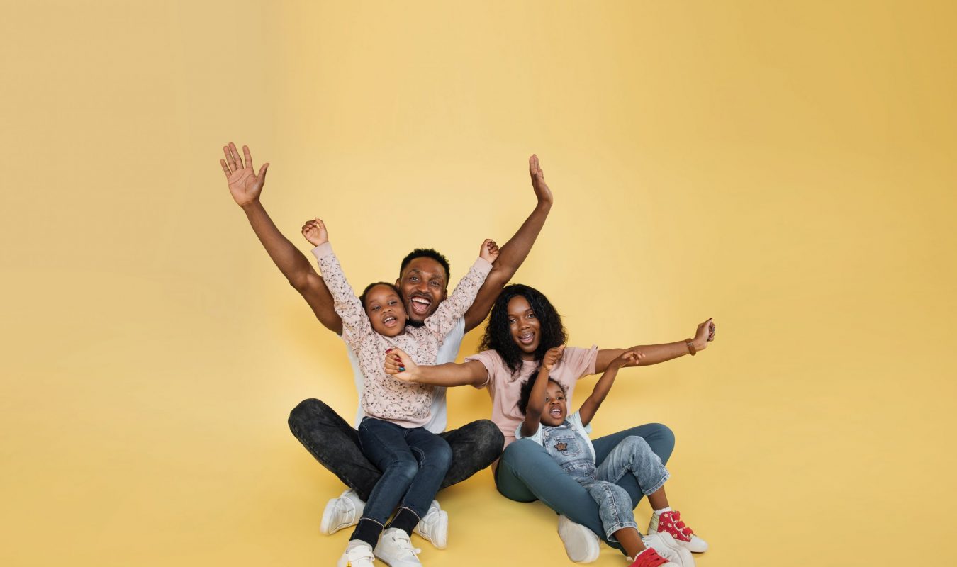 Family fun. Young african american parents spending time with their little daughters with open arms, laughing together over yellow background, banner, panorama, copy space.