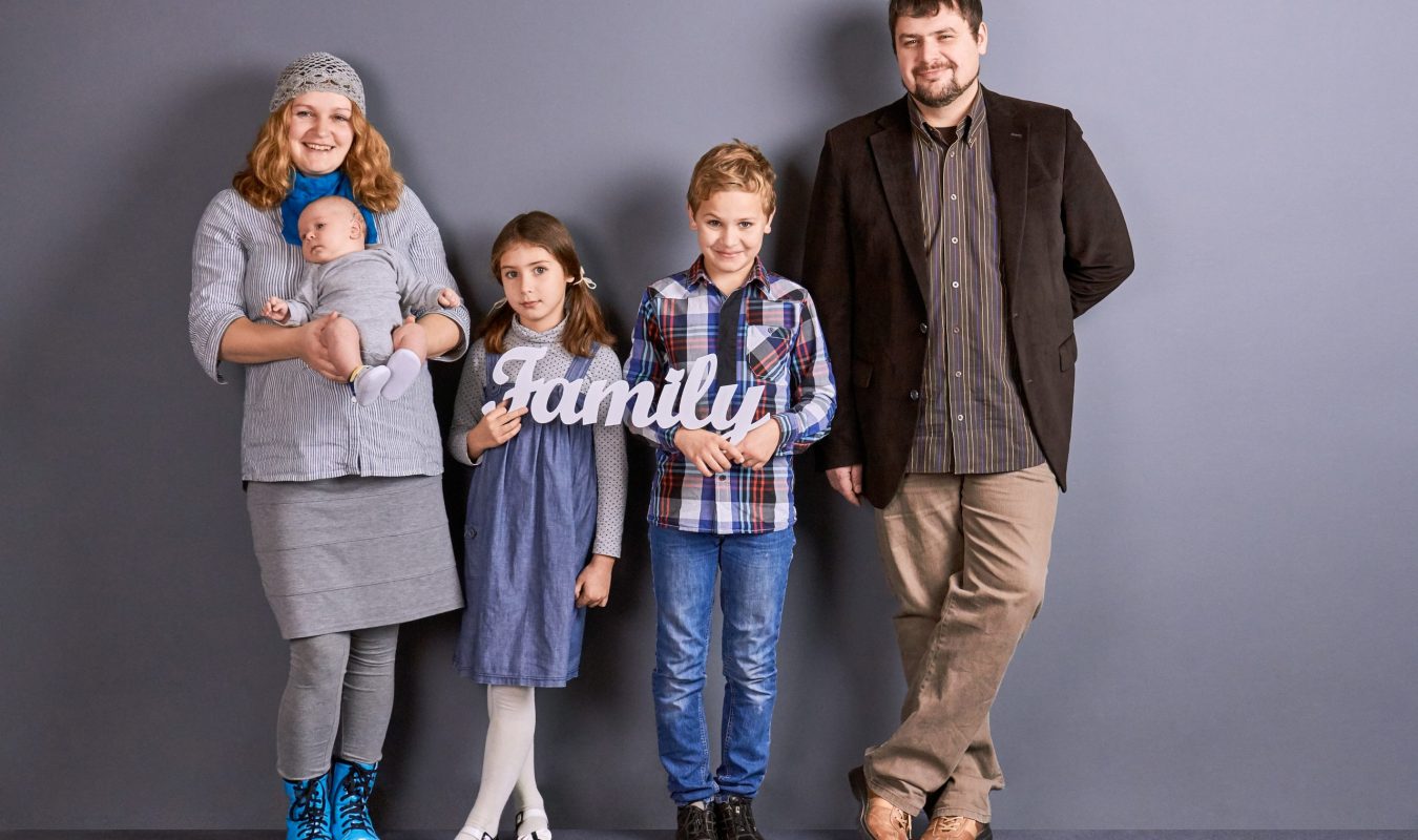 Photo of of beautiful young family. Three cute kids, mother and father standing on grey background. Concept of happy parenting.