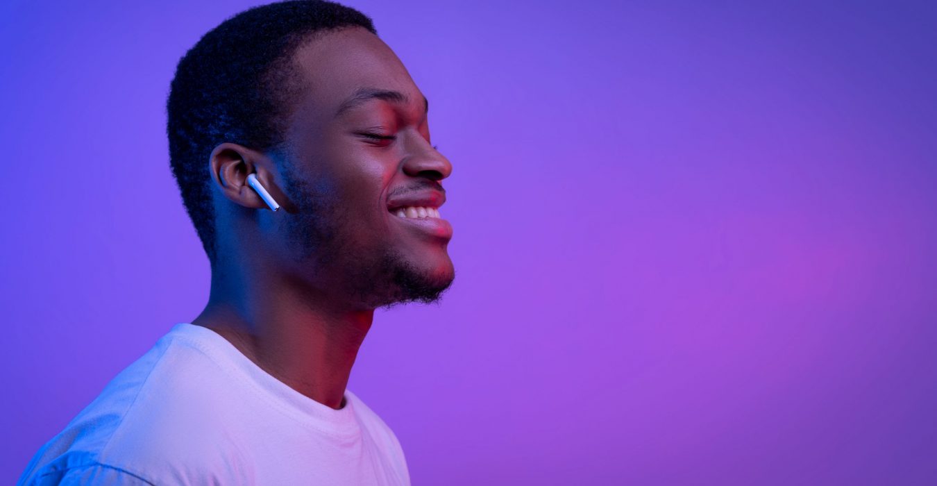 Relaxed African American Man Listening Music In Wireless Airpods Earphones In Neon Light, Smiling Black Guy Wearing Bluetooth Earpods Enjoying Favorite Songs Over Purple Background, Panorama