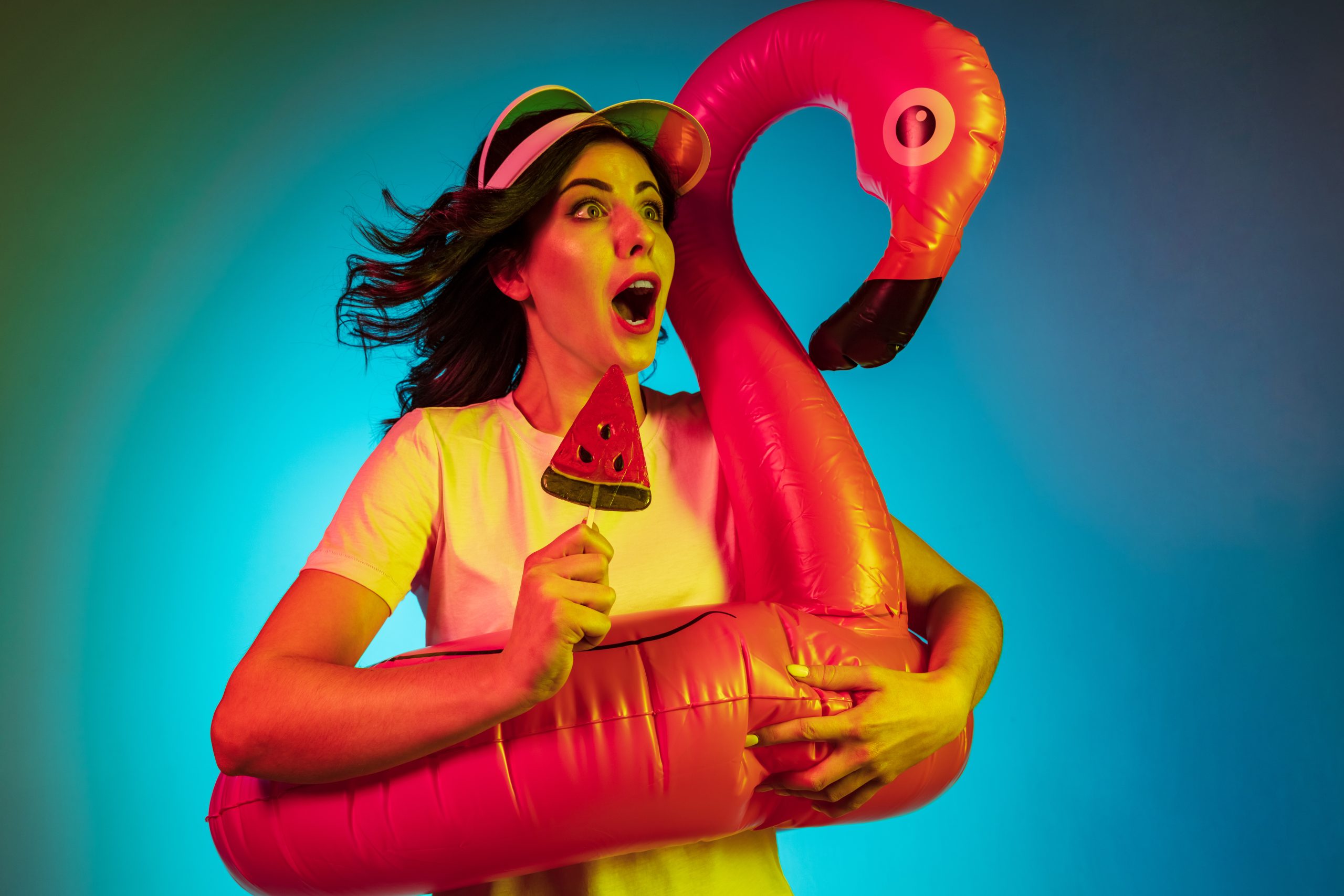 Happy young woman in a cap and in a rubber flamingo with the candy on trendy blue neon studio background. Female portrait. Concept of human emotions, facial expression, summer holidays or weekend.