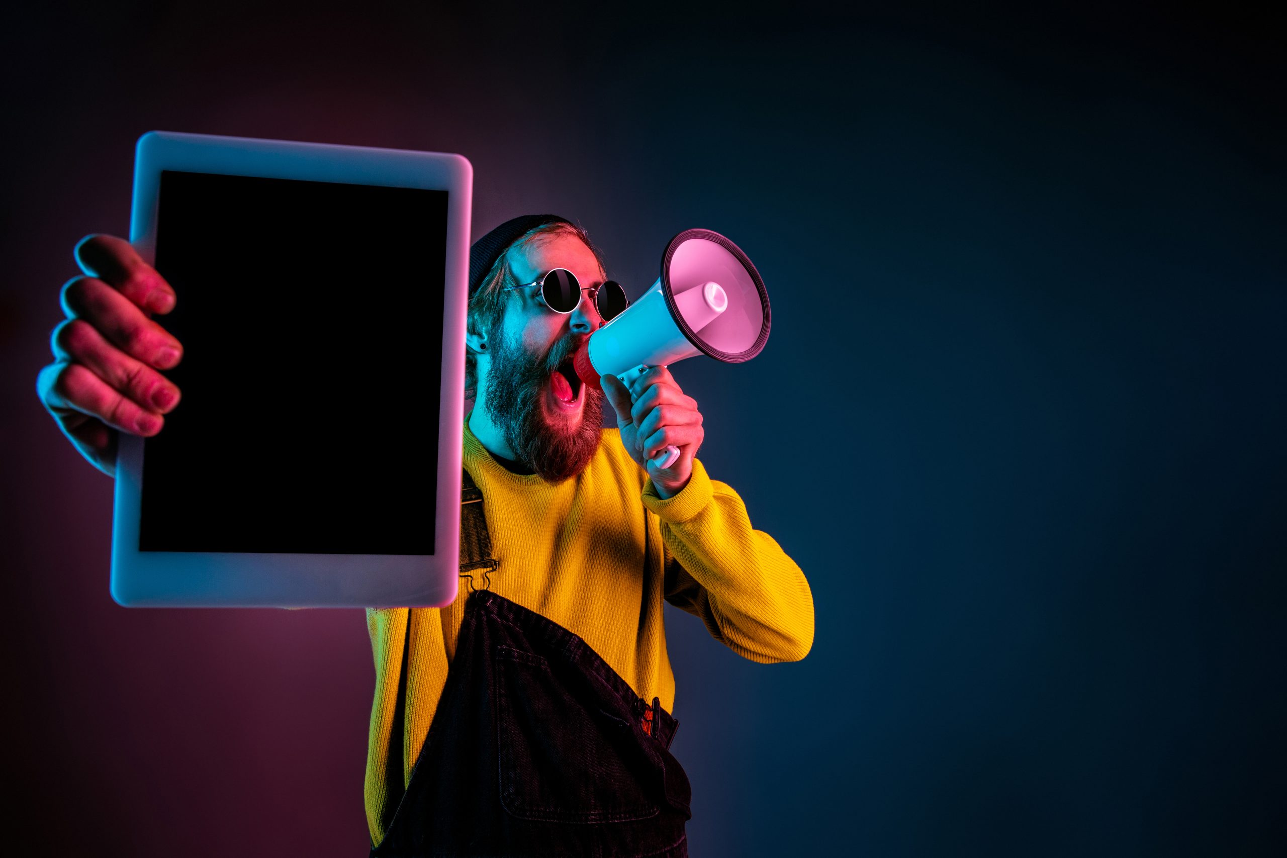 Showing tablet's blank screen. Caucasian man's portrait on gradient studio background in neon light. Beautiful male model with hipster style. Concept of human emotions, facial expression, sales, ad.