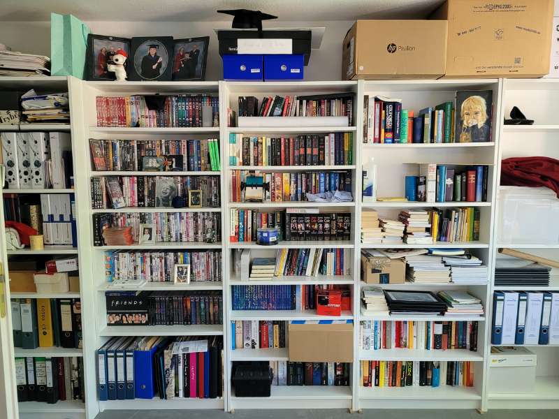 Blick auf fünf Bücherregale, die sehr bunt mit vielen Büchern, DVDs und anderem vollgestopft sind.