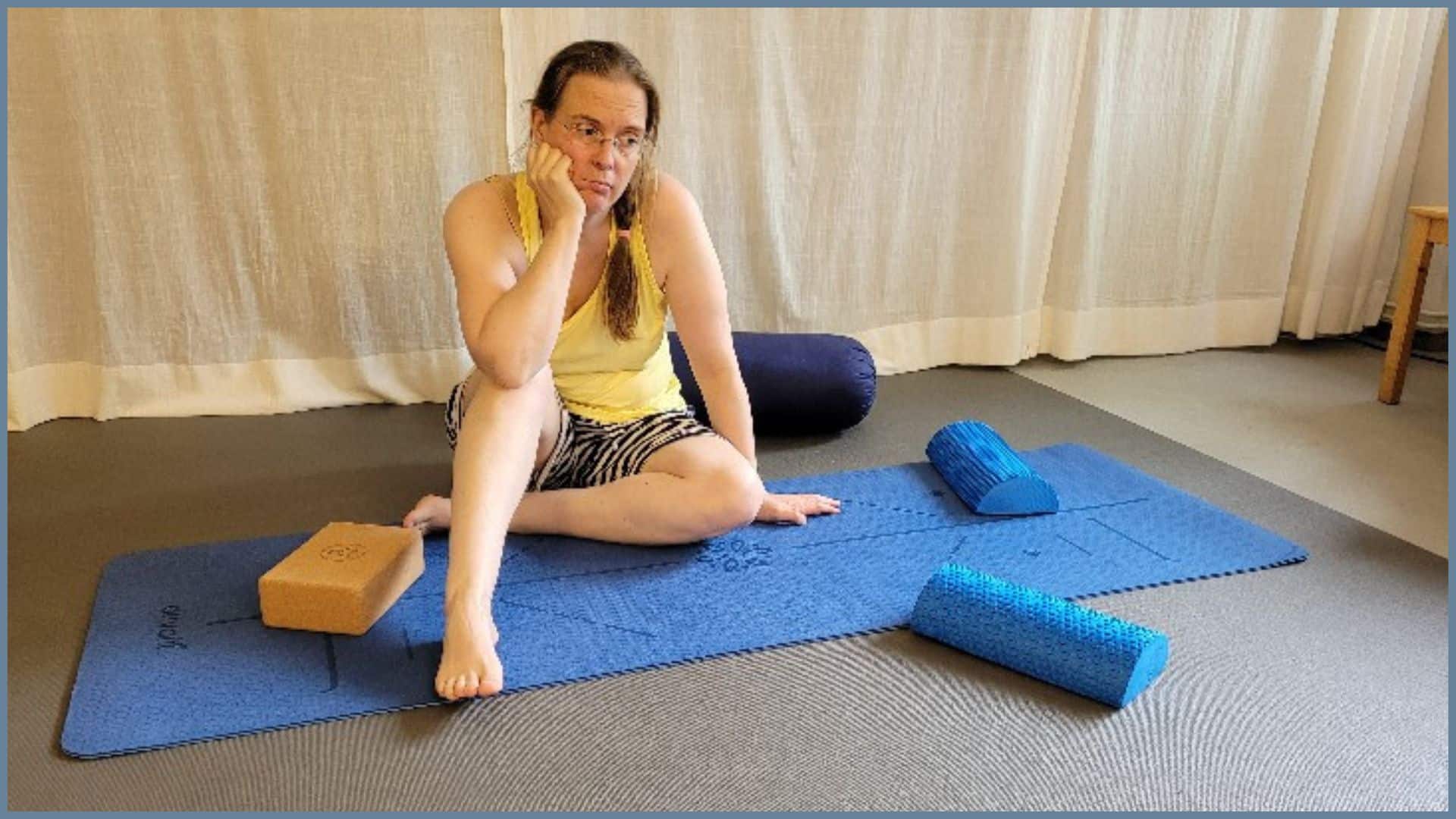 Eine Frau in Sportklamotten sitzt auf einer Yogamatte. Sie stützt ihr Gesicht in eine Hand auf und guckt unzufrieden.