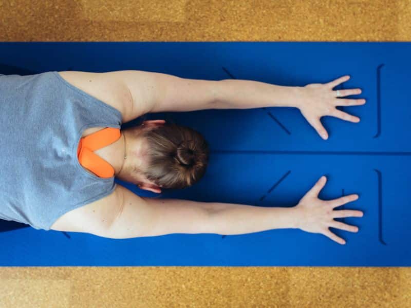 Blick von oben auf eine Yogamatte. Darauf sieht man den Oberkörper einer Frau, die ihre Arme ausgestreckt hat und mit den Handflächen auf die Matte drückt. Man kann nicht erkennen, ob sie auf dem Bauch liegt oder kniet, weil der Teil außerhalb des Bildes ist. Ihr Gesicht zeigt zur Matte, man sieht also nur den Rücken und Hinterkopf und die ausgestreckten Arme. Dies ist eine halb-aktive Pose im Beckenboden-Training, bei der die Schultern gedehnt werden und der Core aktiviert werden kann.