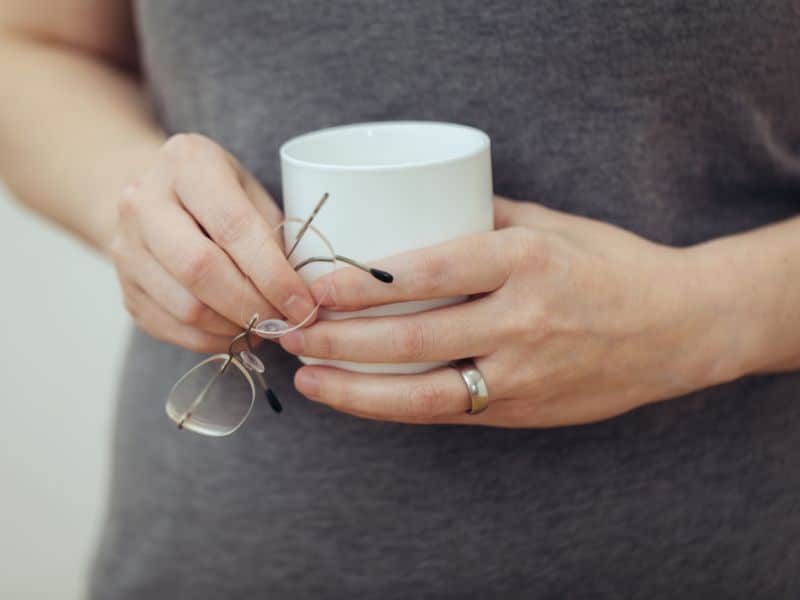 Es sind zwei Frauenhände zu sehen, die vor dem Bauch eine Tasse und eine randlose Brille halten. An der linken Ringfinger steckt ein schlichter silberfarbener Ring.