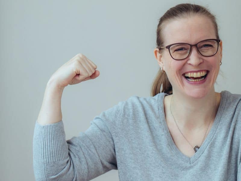 Ein Portrtaitfoto einer herzhaft lachenden Frau. Sie hält ihren rechten Arm so, dass sie ihren Bizeps beugt, die rechte Hand ist dabei zur Faust geballt. Die Pose stellt eine übliche Haltung zur Demonstration von Kraft im Arm dar.