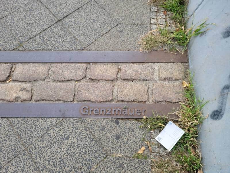 Auf dem Straßenboden ist mit zweireihig verlegten Pflastersteinen umrahmt von Metallschienen der Verlauf der alten Grenze zwischen Ost- und West-Berlin markiert. Auf dem einen Metallstreifen ist das Wort Grenzmauer erhaben eingearbeitet.