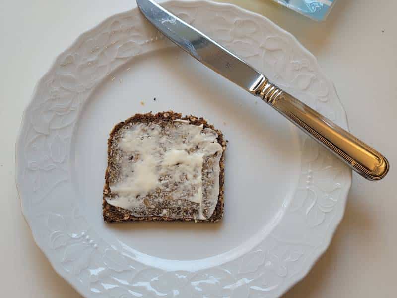 Eine Scheibe Brot mit Butter auf einem weißen Teller. Diagonal über die Tellerecke liegt ein Messer.